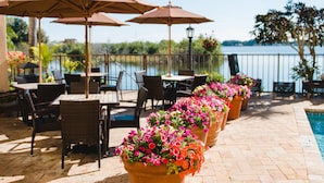An outdoor patio with tables and chairs, surrounded by potted plants and umbrellas, overlooking a body of water. - Inn On The Lakes (Sebring)