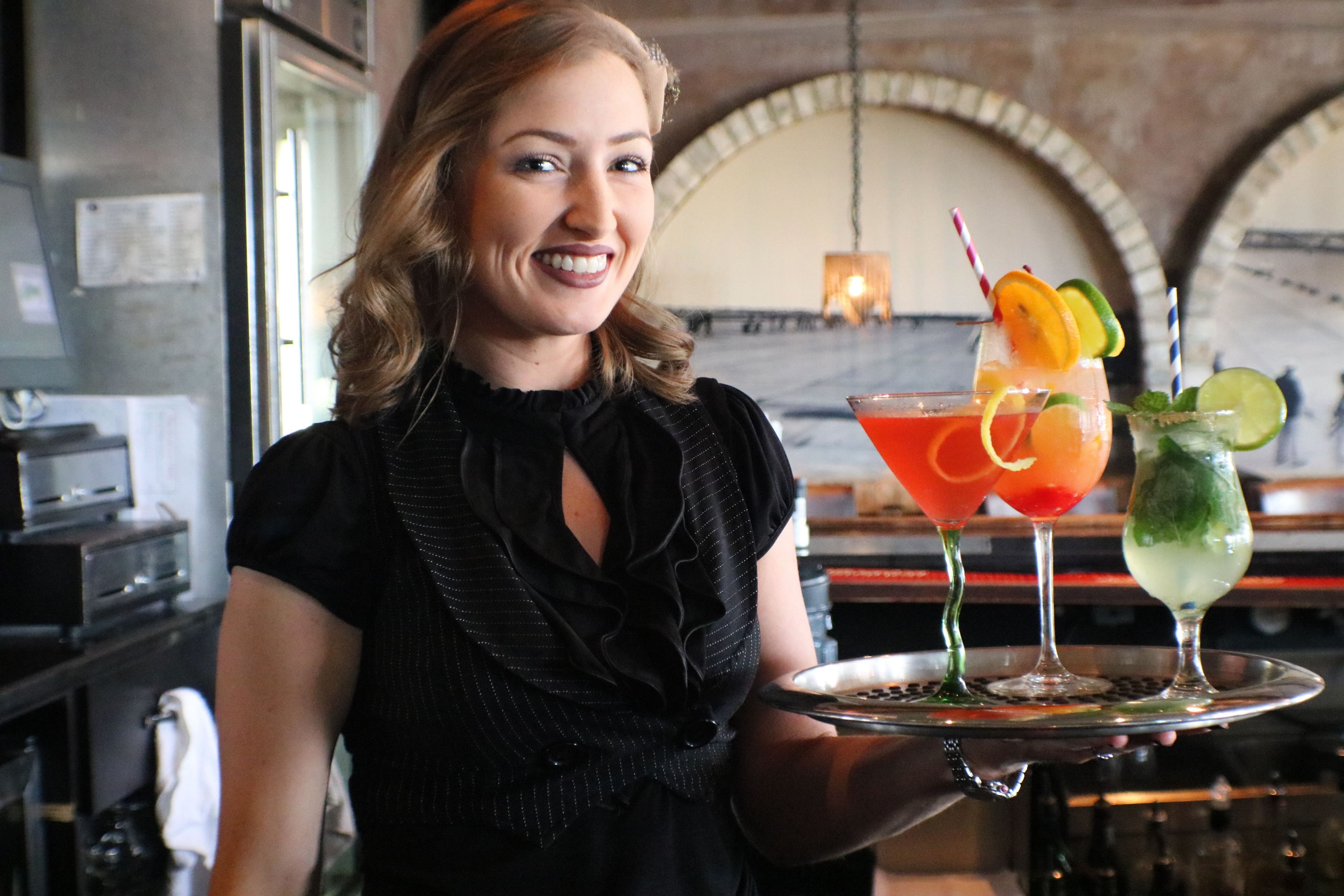A person holding a tray with three drinks, a kitchen area with appliances, and a brick wall with an arched window.