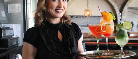 A person holding a tray with three drinks, a kitchen area with appliances, and a brick wall with an arched window.