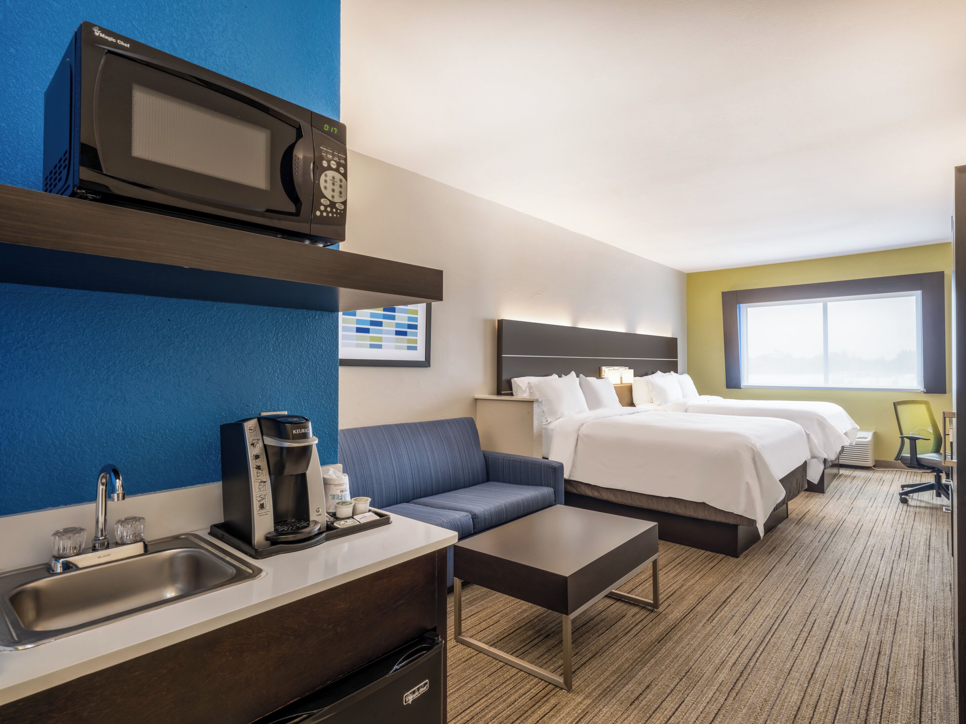 A hotel room with a kitchenette, a bed, a sofa, a coffee maker, and a window.