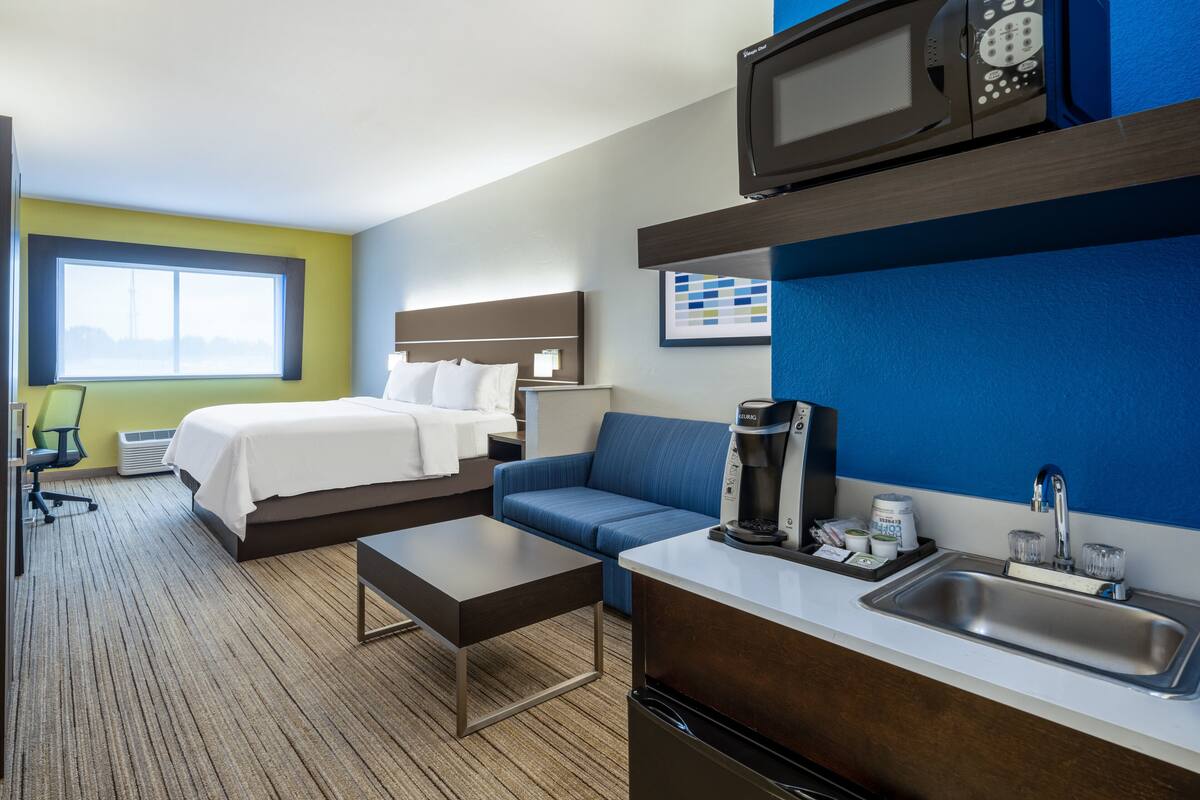 A hotel room with a bed, a sofa, a coffee machine, a microwave, a sink, and a window with a view.