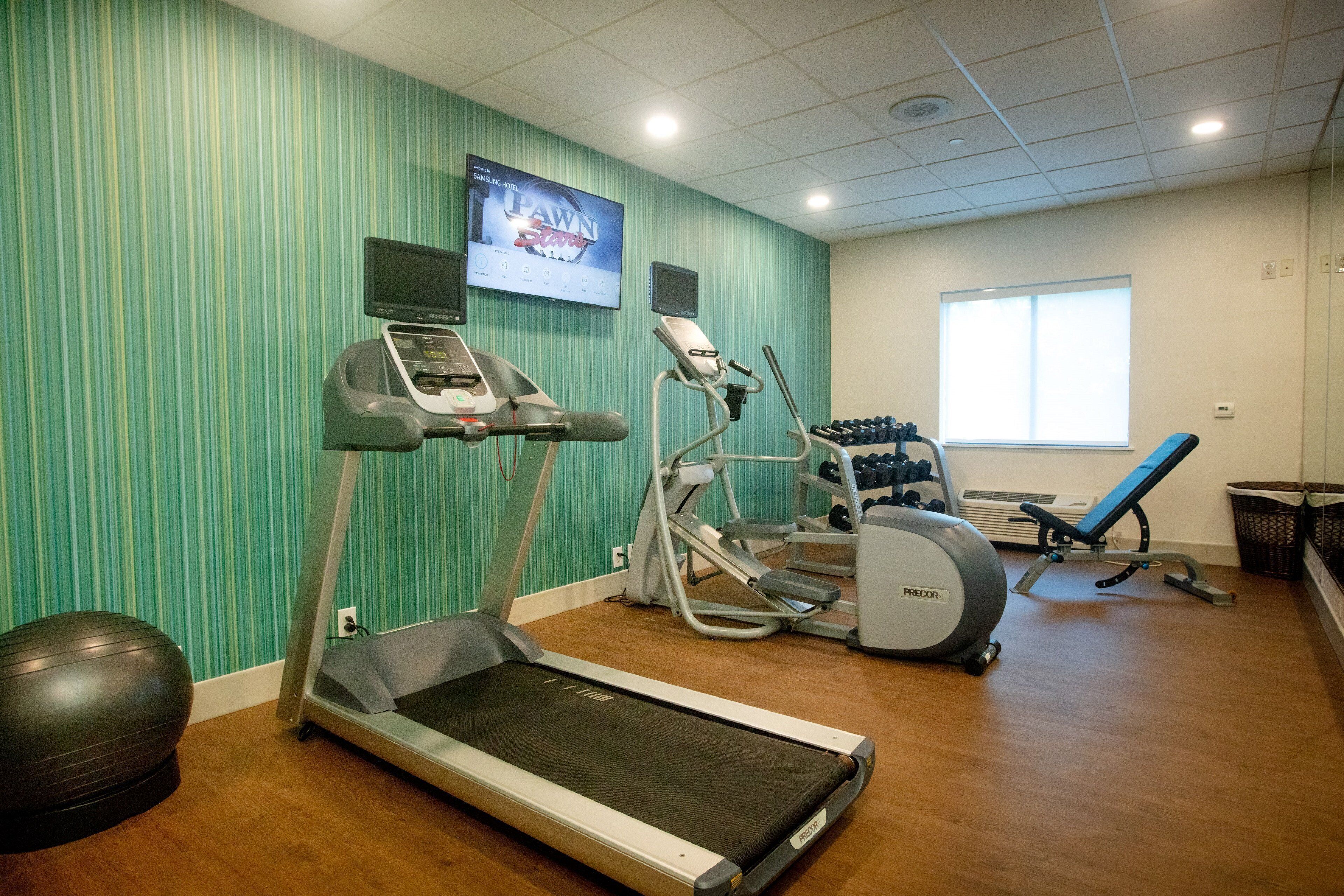 A gym with a treadmill, elliptical, and weights, a TV on the wall, and a window.