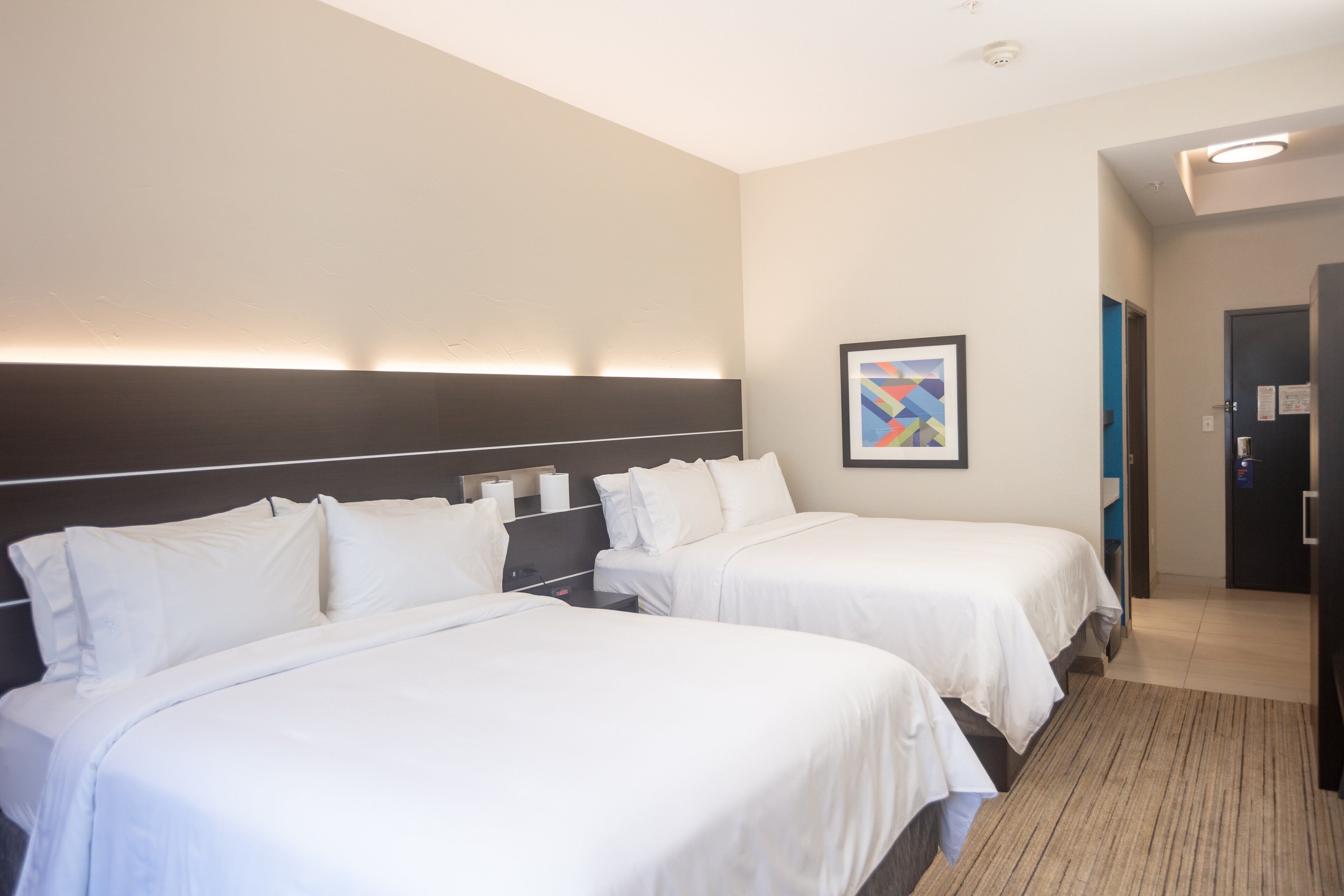 Habitación de hotel con dos camas, cabecero, un cuadro en la pared y una puerta que da a otra habitación.