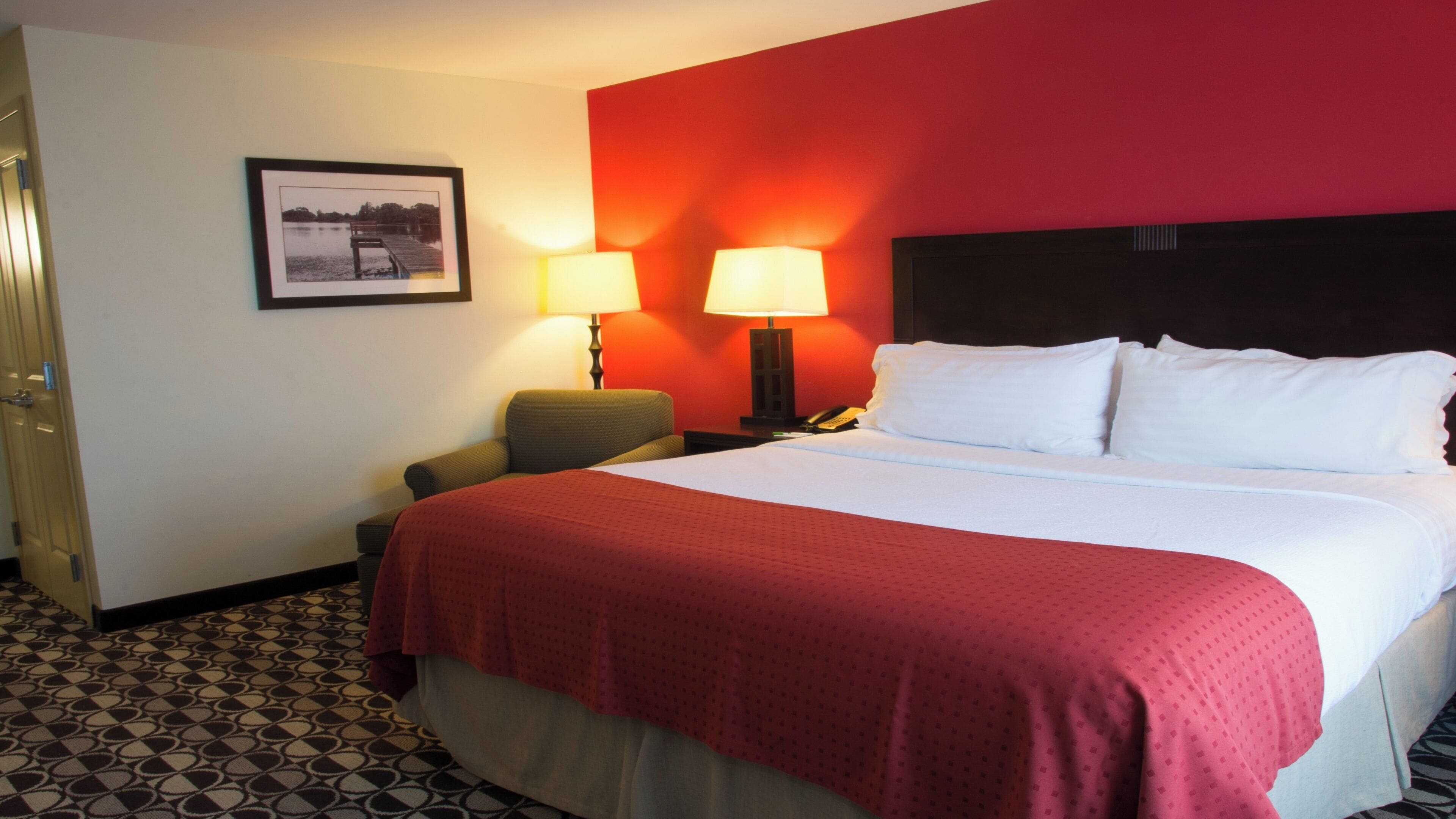 A hotel room with a large bed, two bedside lamps, a chair, a desk with a telephone, and a framed picture on the wall.
