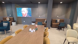 Une salle de conférence avec une longue table, des chaises jaunes et un grand écran sur lequel est affiché un portrait.