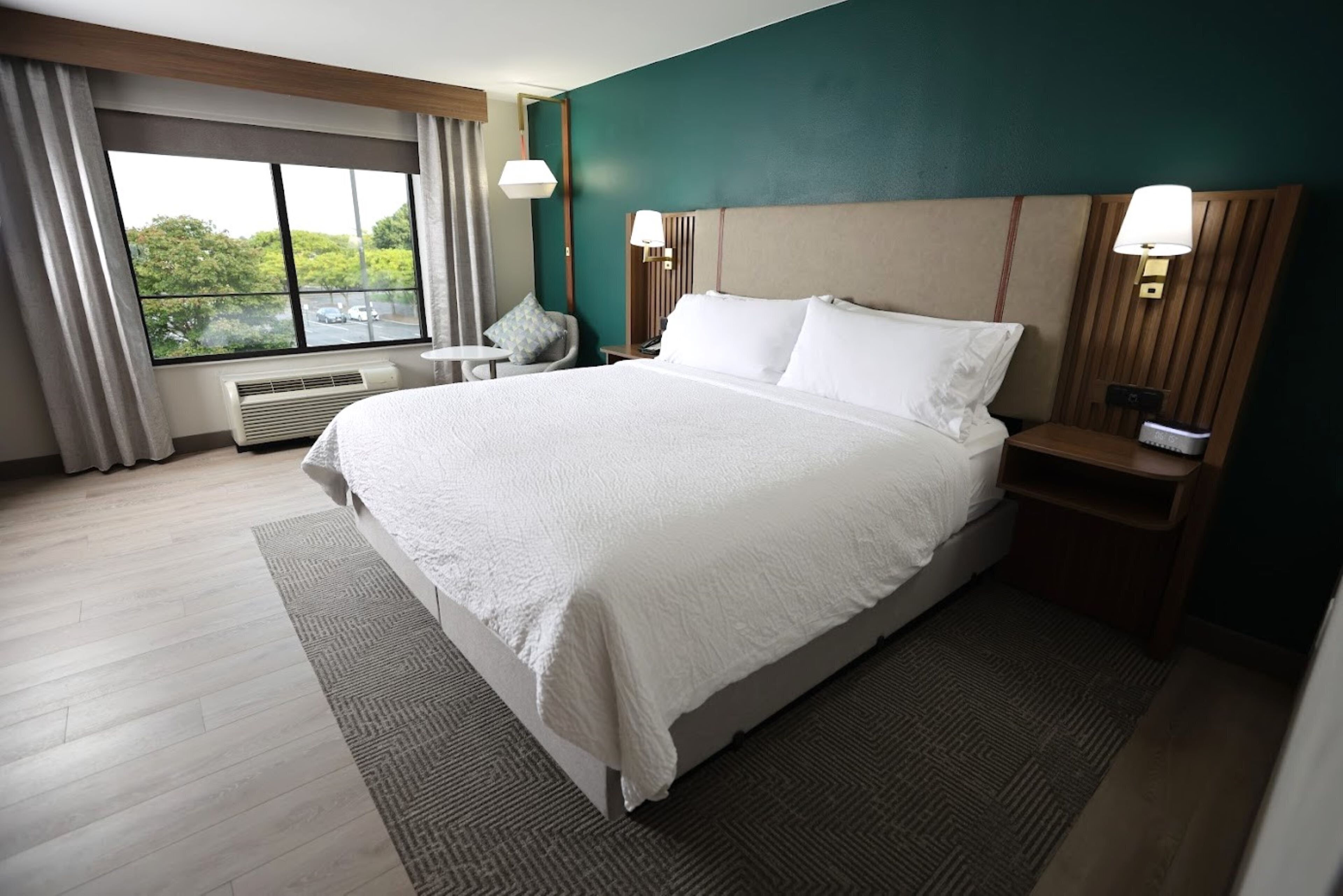 A hotel room with a large bed, a chair, a nightstand, a lamp, and a window with curtains.