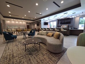 A modern lounge area with a curved sofa, armchairs, and a coffee table. There is a bar area with a TV and bar stools. - Holiday Inn St. Louis Fairview Heights by IHG (Fairview Heights)