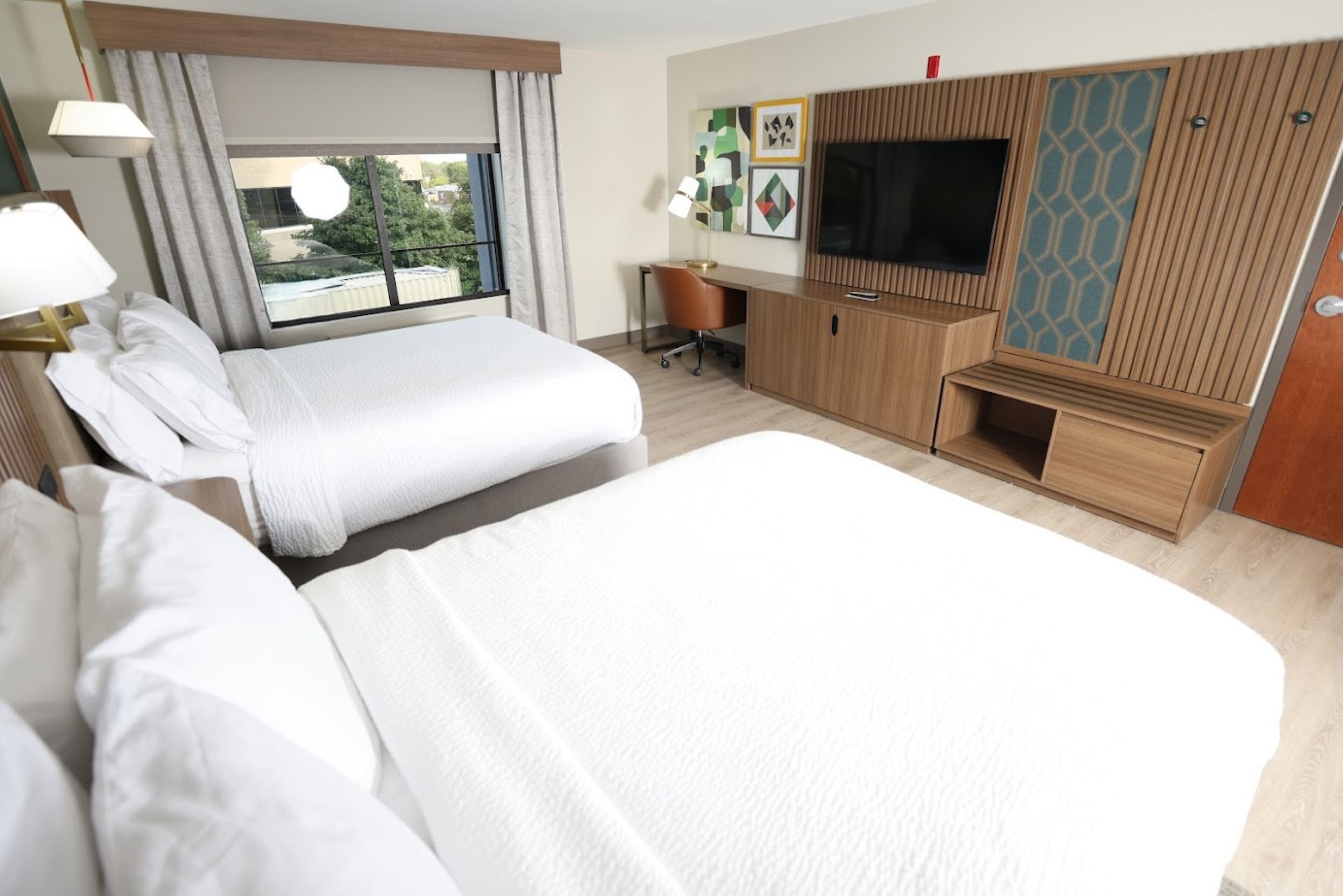 A hotel room with two beds, a desk, a TV, and a window with a view of greenery.