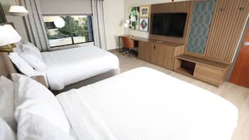 A hotel room with two beds, a desk, a TV, and a window with a view of greenery.