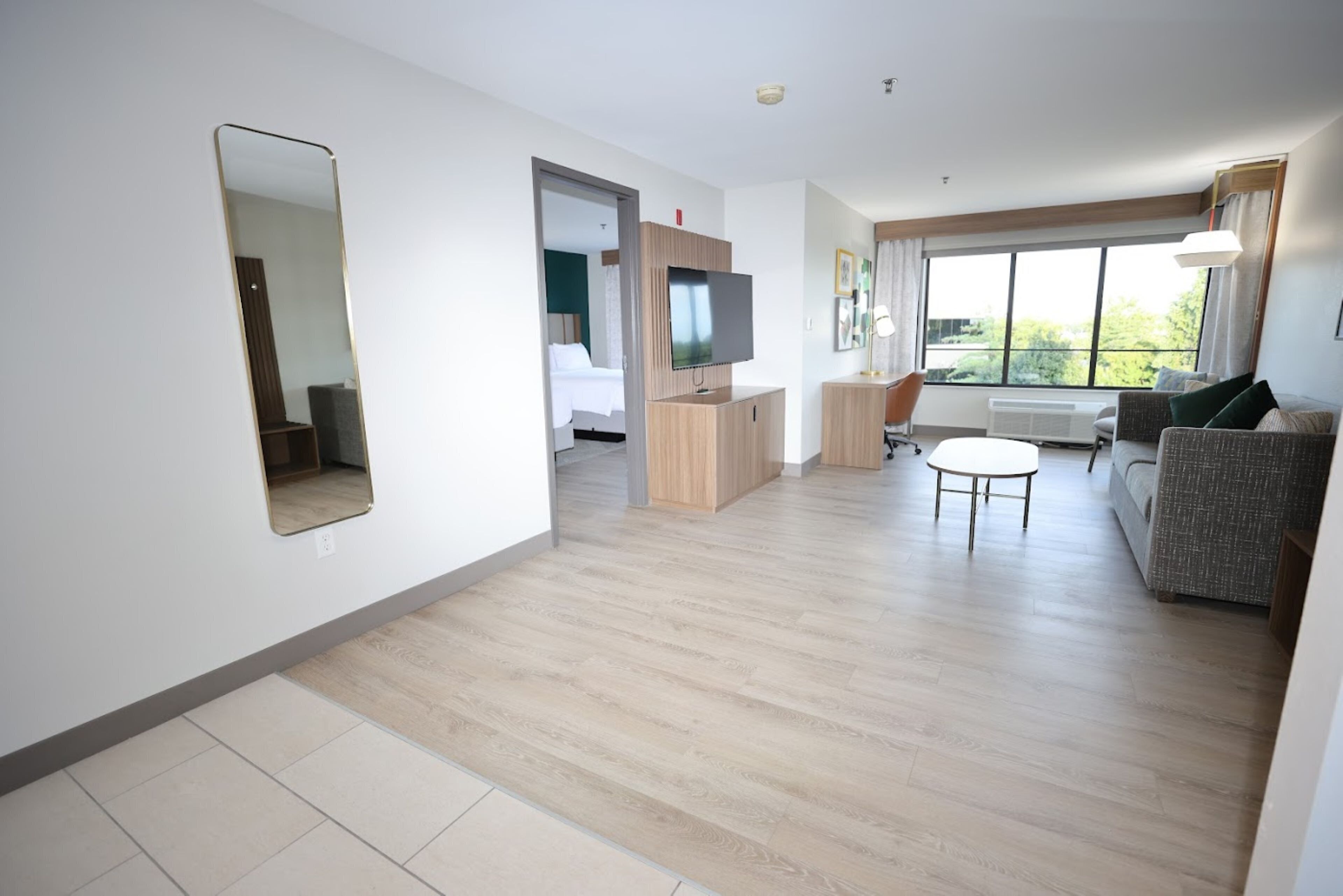 A modern hotel room with a large mirror, a television, a sofa, a small table, and a window with a view of greenery.