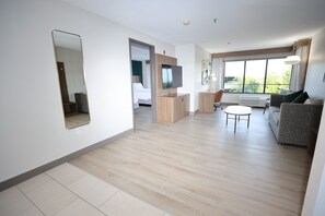 A modern hotel room with a large mirror, a television, a sofa, a small table, and a window with a view of greenery. - Holiday Inn St. Louis Fairview Heights by IHG (Fairview Heights)