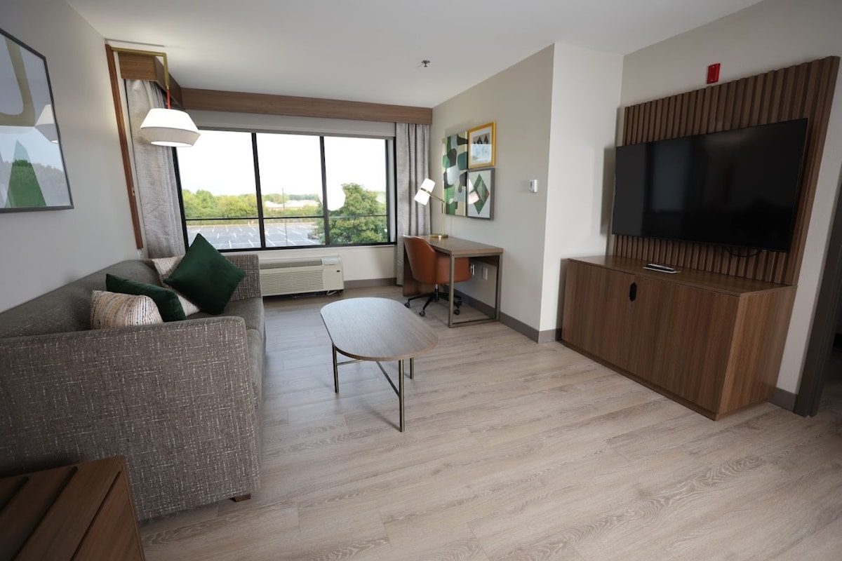 Una sala moderna con sofá, mesa de comedor, televisor y un amplio ventanal con vistas a zonas verdes.