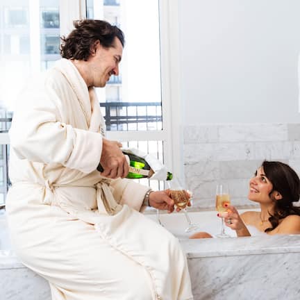 A man in a white bathrobe pouring champagne into a glass held by a woman in a bathtub.
