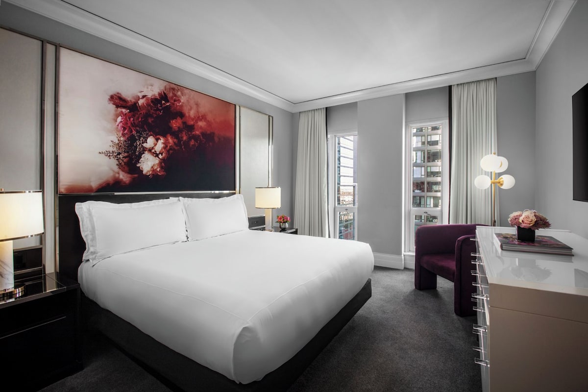 A modern hotel room with a large bed, a desk, a chair, and a large abstract painting on the wall.