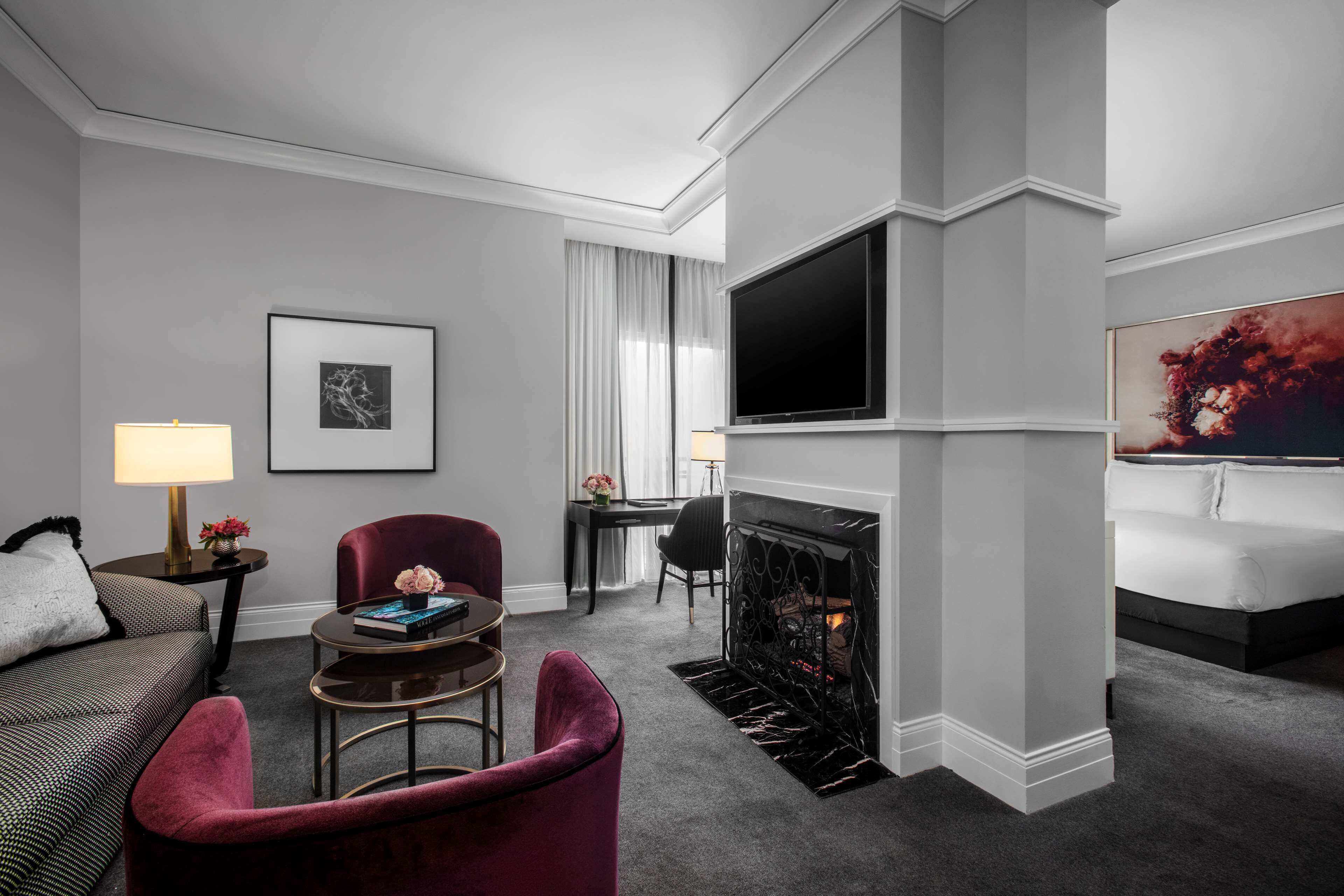 Una habitación de hotel moderna con chimenea, un televisor de pantalla plana montado encima, un sofá cómodo, un sillón rojo y una mesita con una lámpara y objetos decorativos.