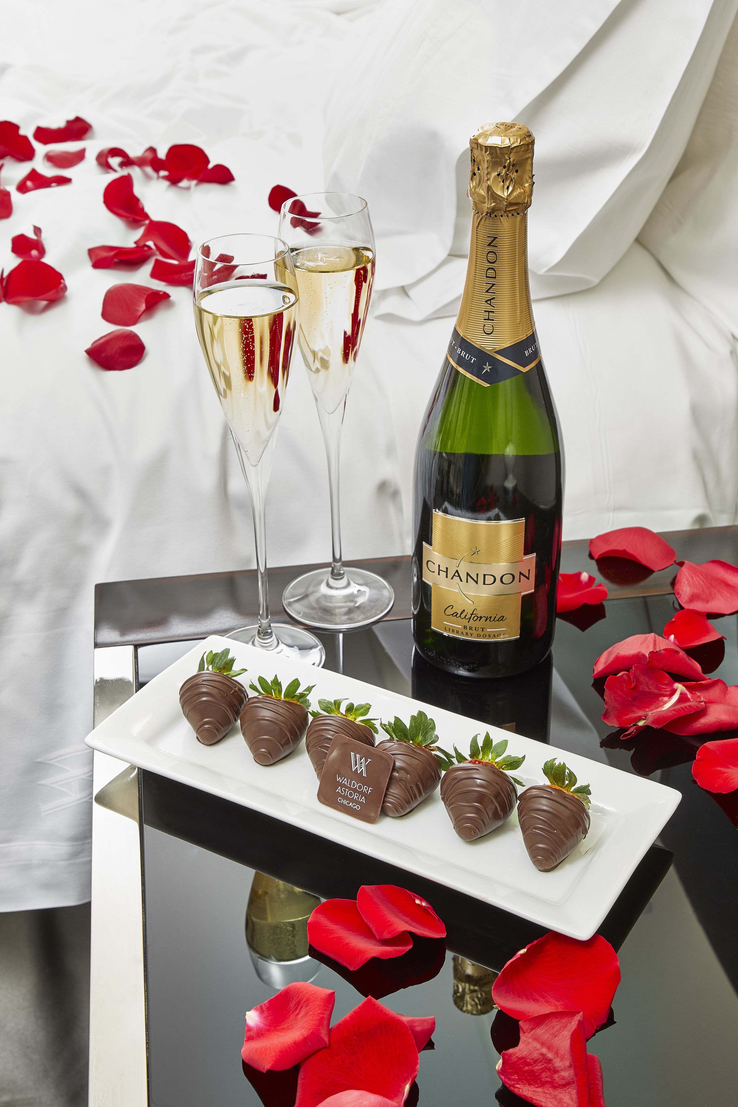 A bottle of Chandon California Brut champagne, two glasses of champagne, and chocolate-covered strawberries on a reflective surface with red rose petals scattered around.