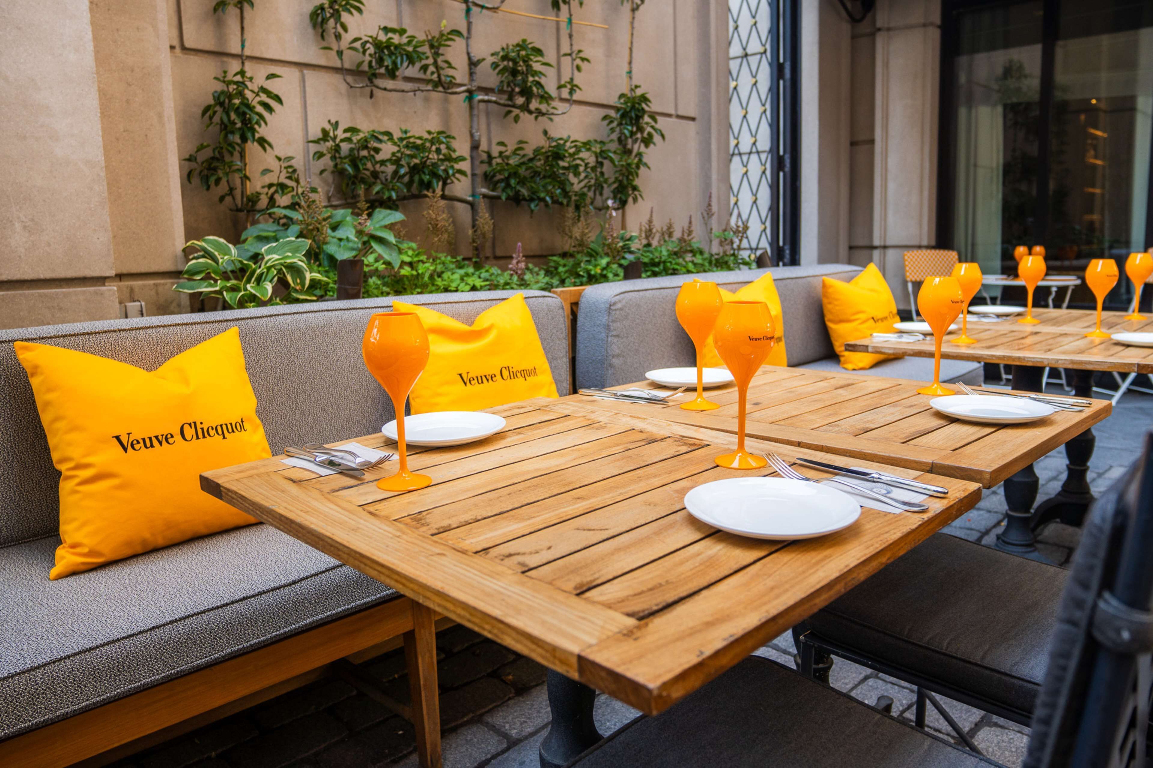 Mesas para comer al aire libre con cojines amarillos y vasos naranjas, frente a un fondo de vegetación y una pared de edificio.