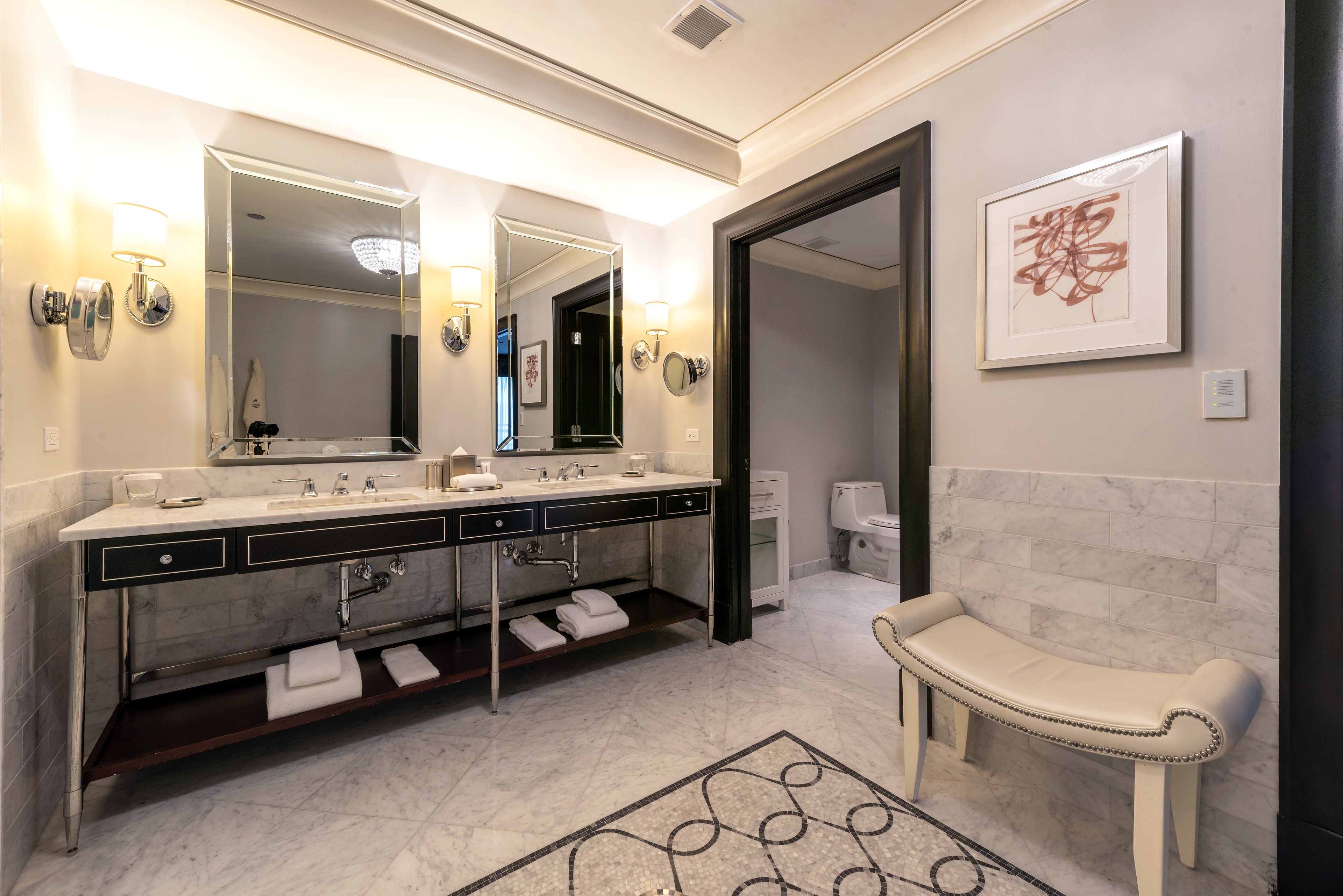 A modern bathroom with a double sink vanity, a large mirror, wall sconces, a bench, and a framed artwork.