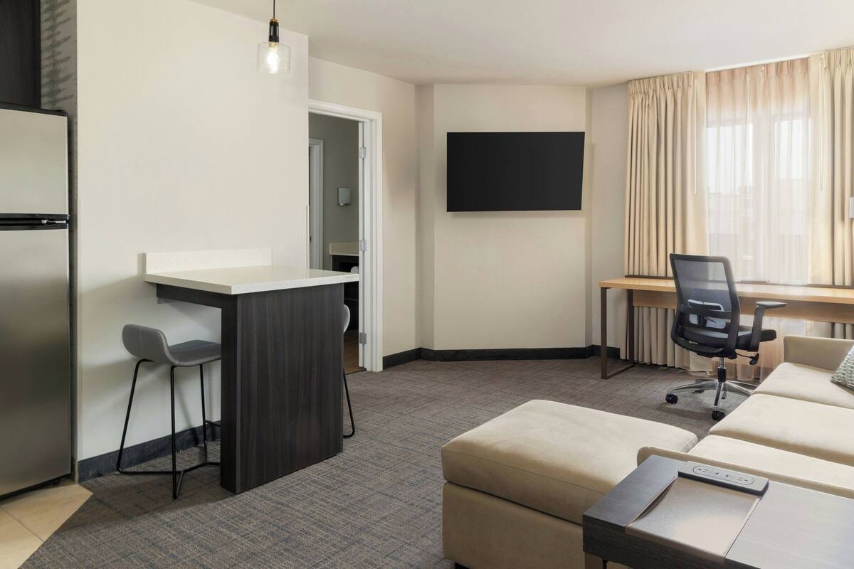 A modern hotel room with a kitchenette, a flat-screen TV, a sofa, and a desk with a chair.