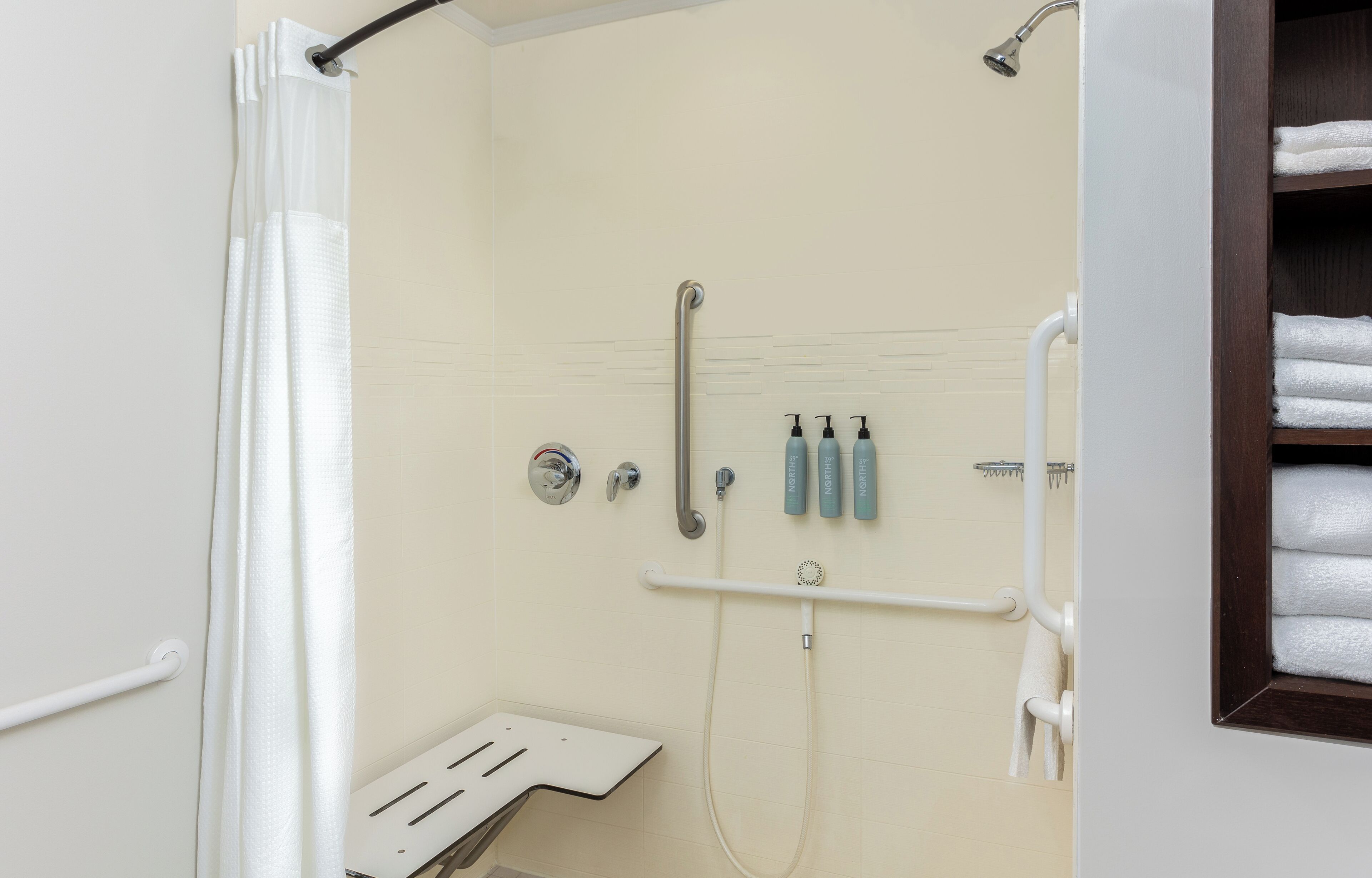 A shower area with a white shower curtain, a grab bar, a handheld showerhead, and a shelf with toiletries.