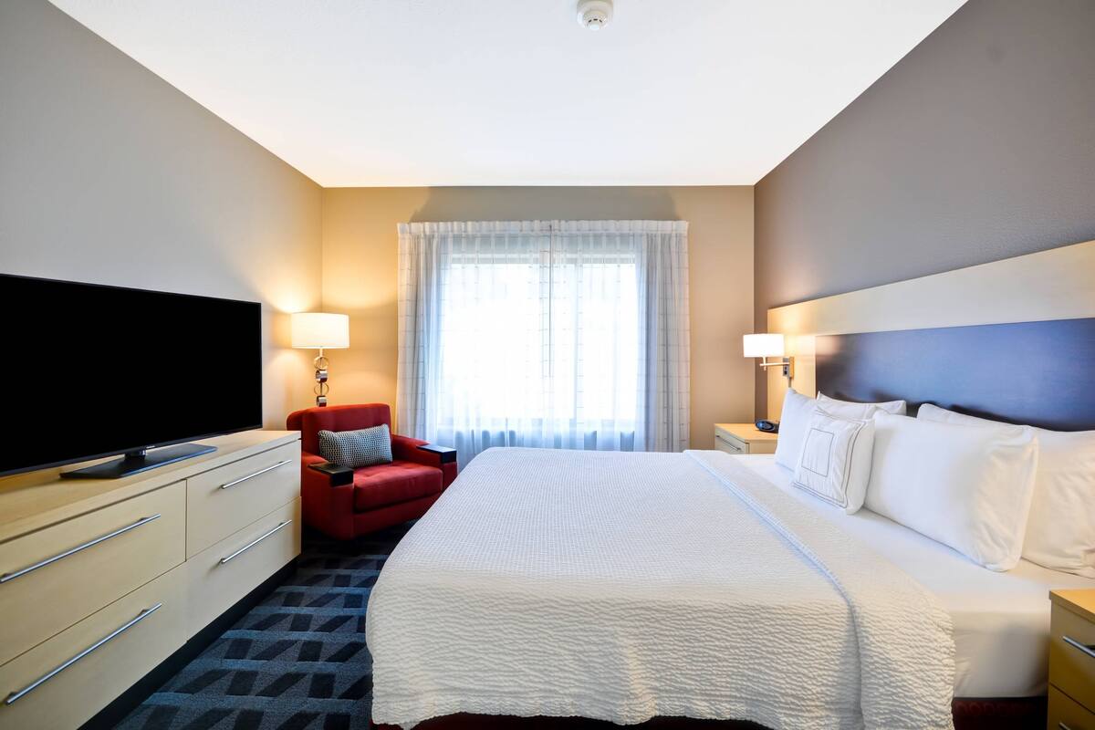 Una habitación de hotel con una cama grande, un televisor en una cómoda, un sillón rojo y una ventana con cortinas.
