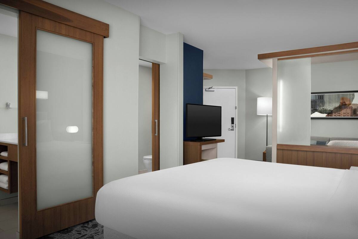A hotel room with a large bed, a TV, a bathroom with a toilet, and a mirror.