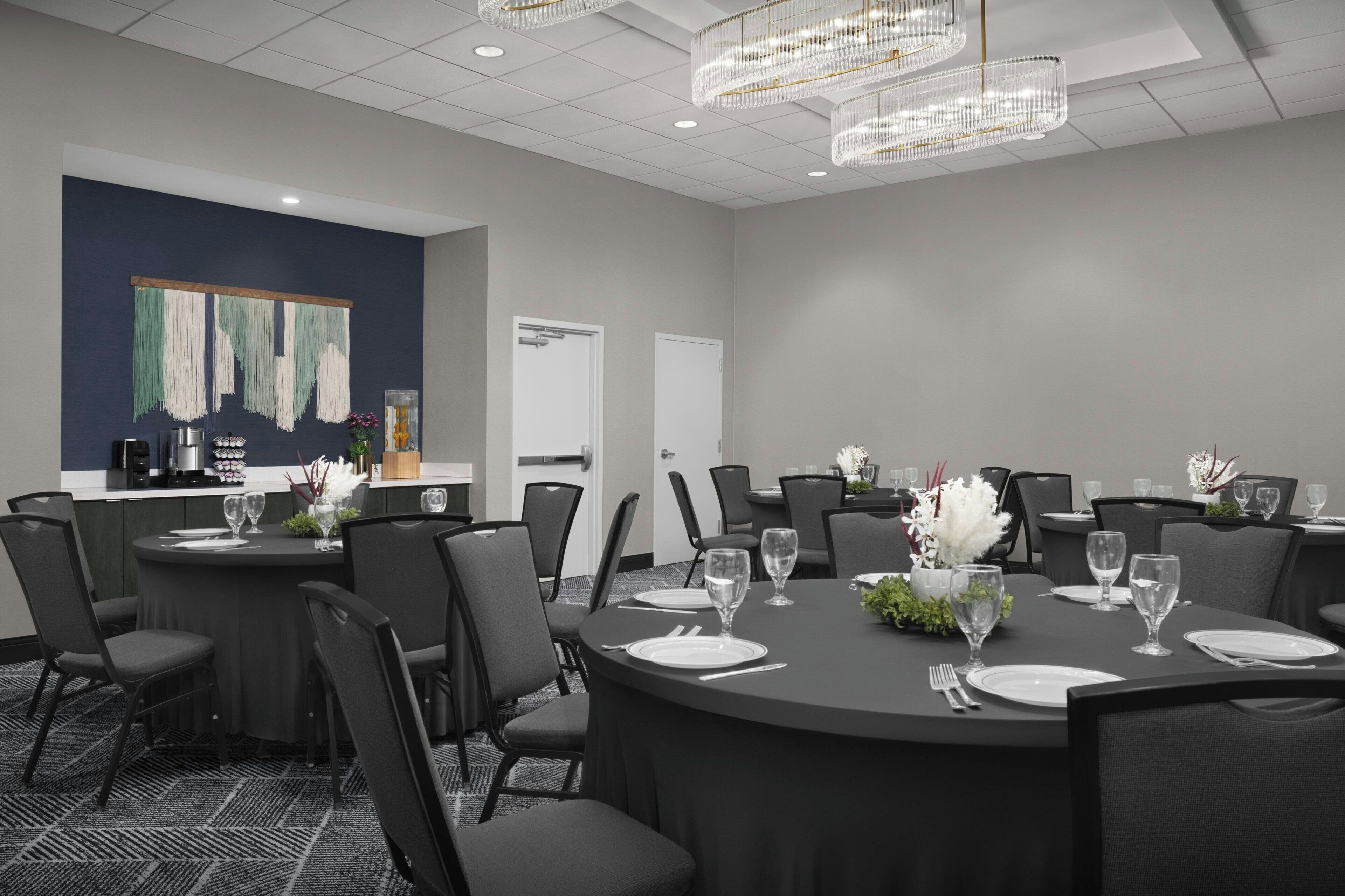 A conference room with round tables, black chairs, and a bar area with a refrigerator and coffee machine.
