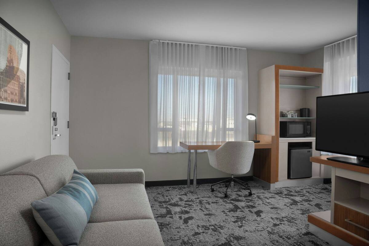 A hotel room with a sofa, a desk, a chair, a television, a microwave, and a refrigerator.