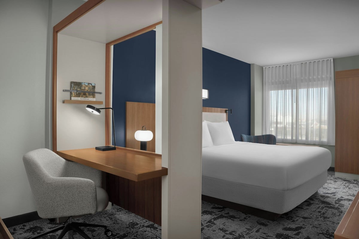 A hotel room with a wooden desk, a chair, a bed, a nightstand, a lamp, and a window with curtains.