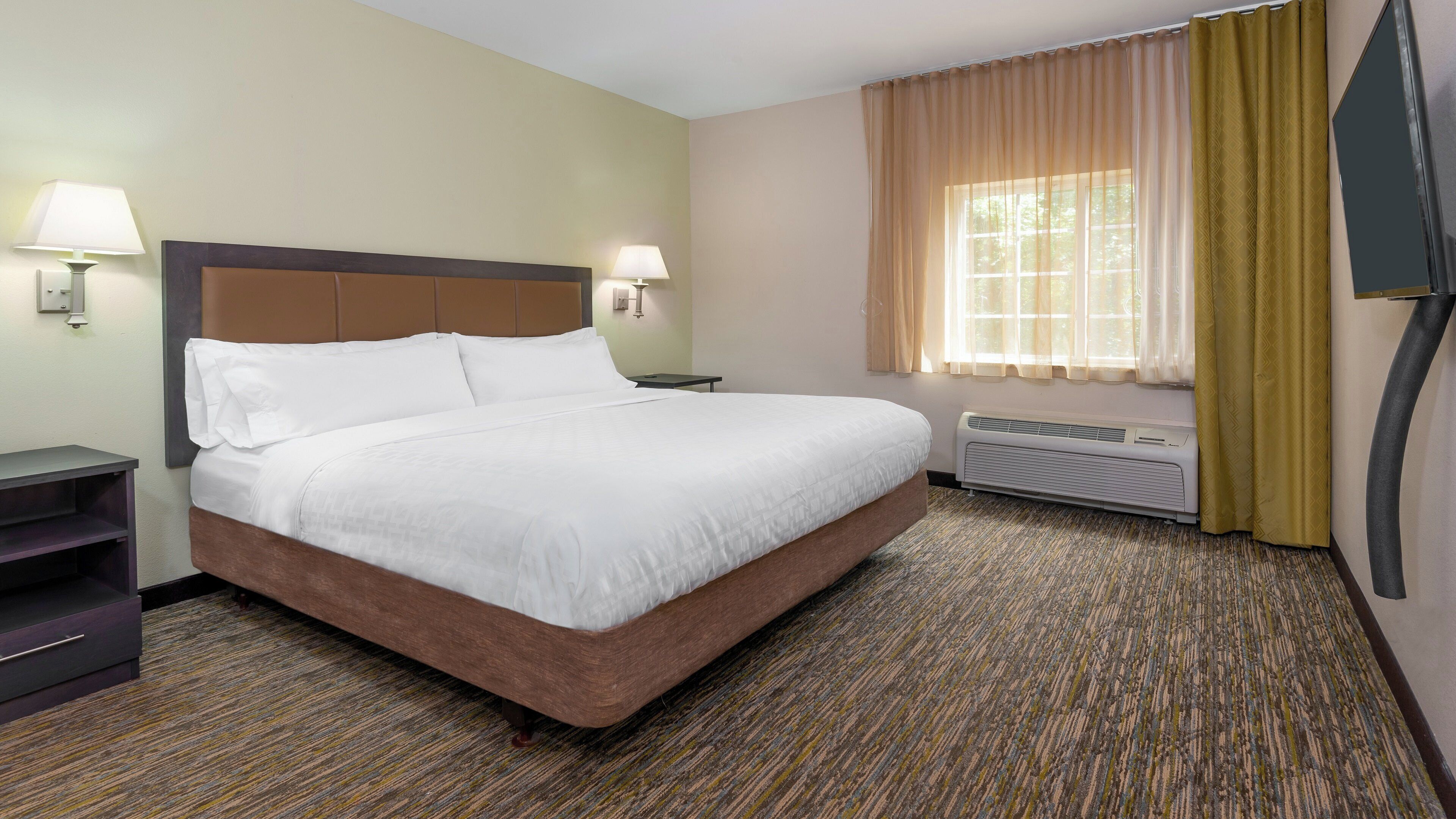 A hotel room with a large bed, two bedside lamps, a window with curtains, a wall-mounted TV, and a heater.
