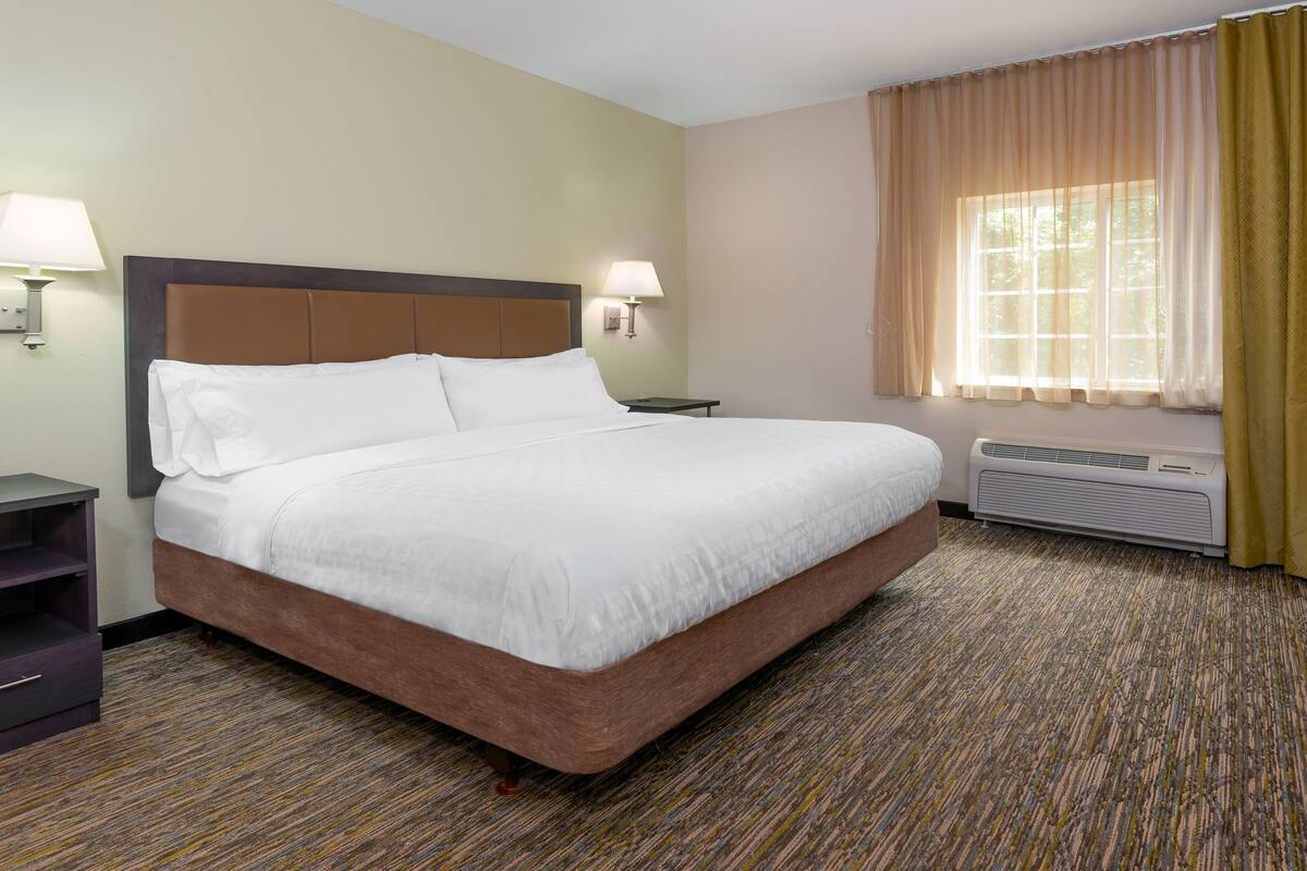 A hotel room with a large bed, two bedside lamps, a window with curtains, a flat-screen TV mounted on the wall, and a built-in air conditioning unit.