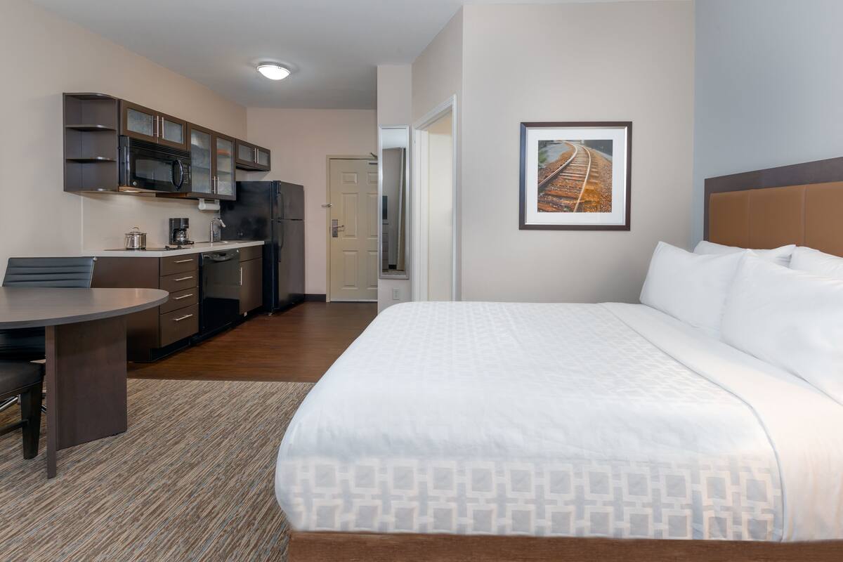 A hotel room with a bed, a desk, a kitchenette, and a framed picture on the wall.