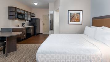 A hotel room with a bed, a desk, a kitchenette, and a framed picture on the wall.