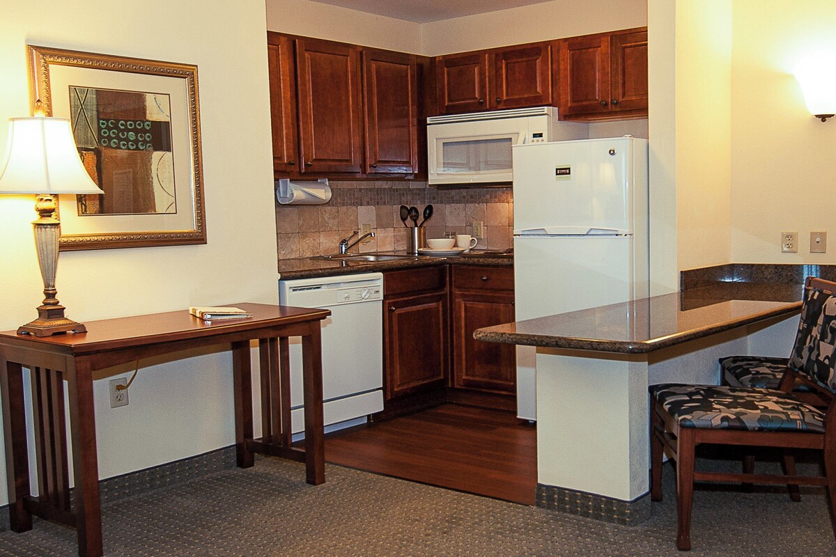 A compact kitchen with wooden cabinets, a microwave, a refrigerator, a sink, and a chair.