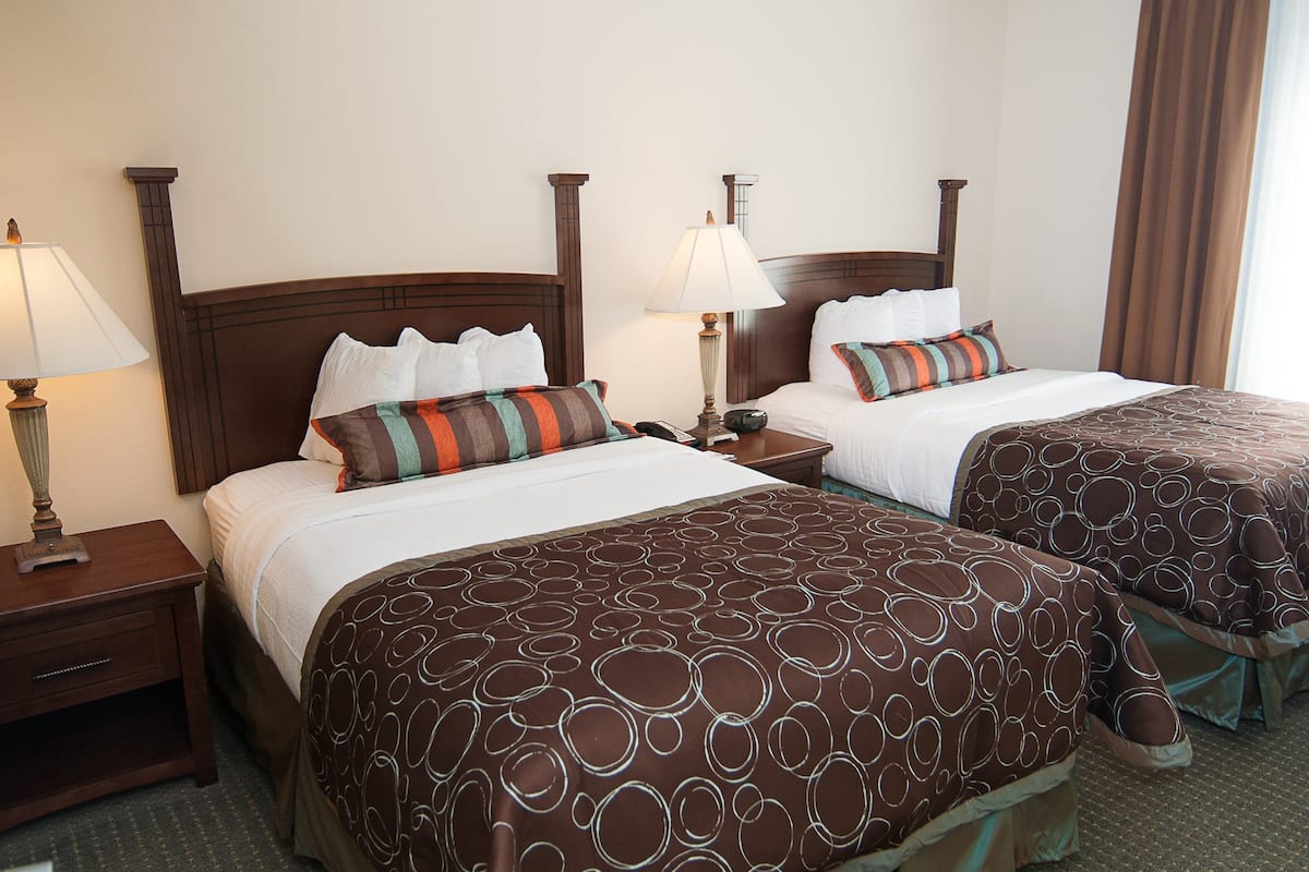 A hotel room with two beds, each with a wooden headboard, a nightstand with a lamp, and a window with curtains.