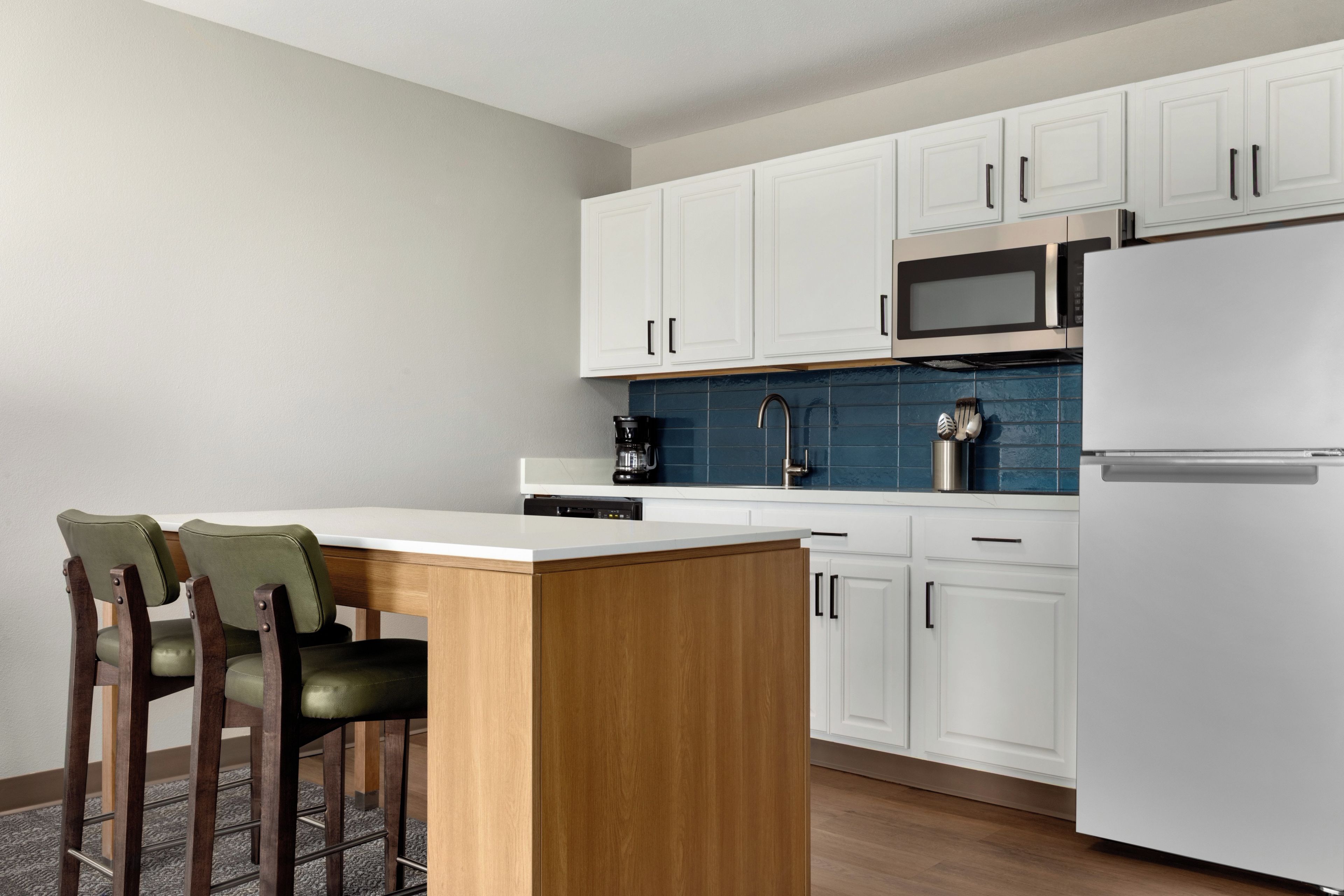A modern kitchen with white cabinets, a stainless steel refrigerator, a microwave, and a coffee maker. There is a wooden island with a bar stool and a blue tiled backsplash.