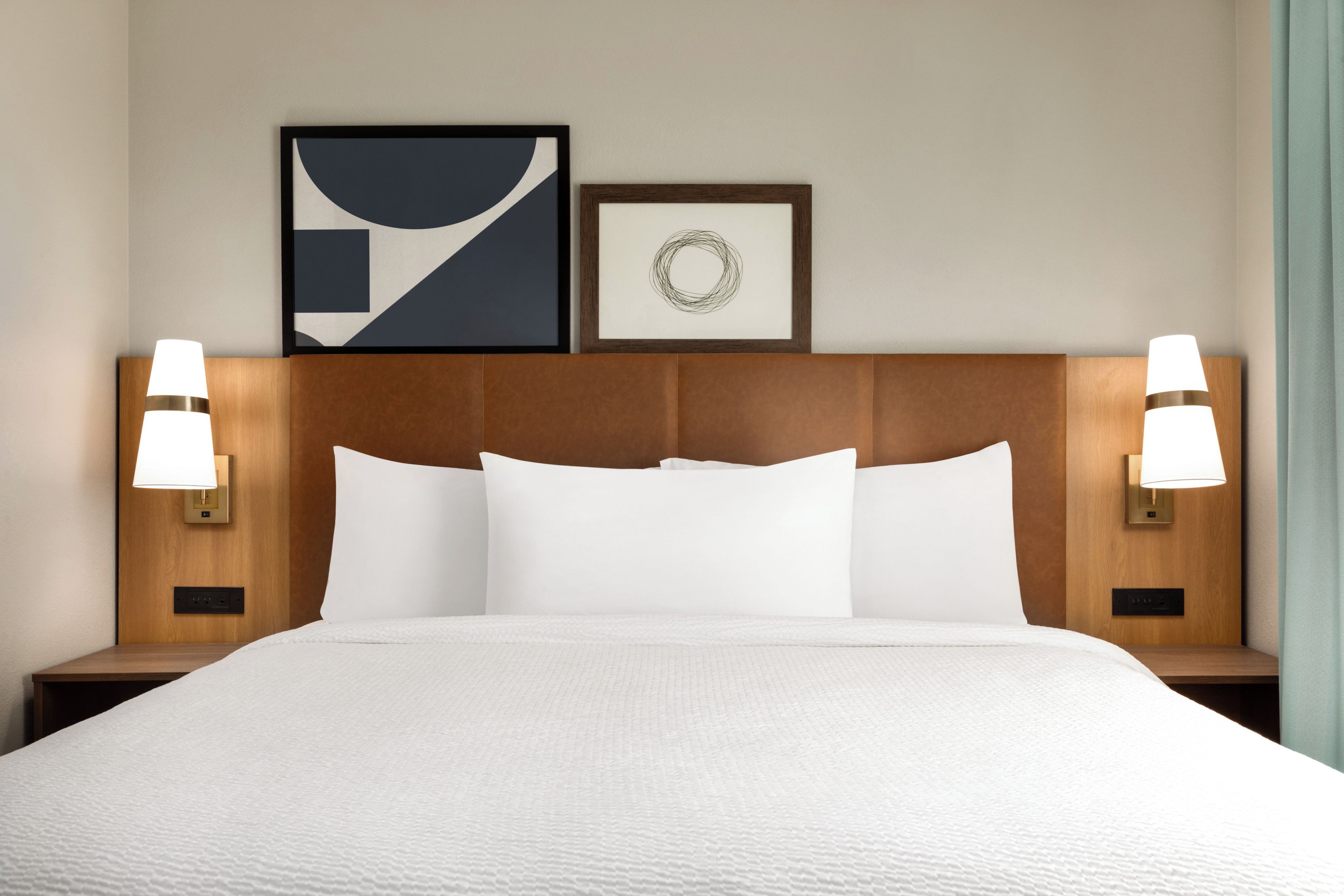 A hotel room with a large bed, two bedside lamps, and two framed artworks on the wall.
