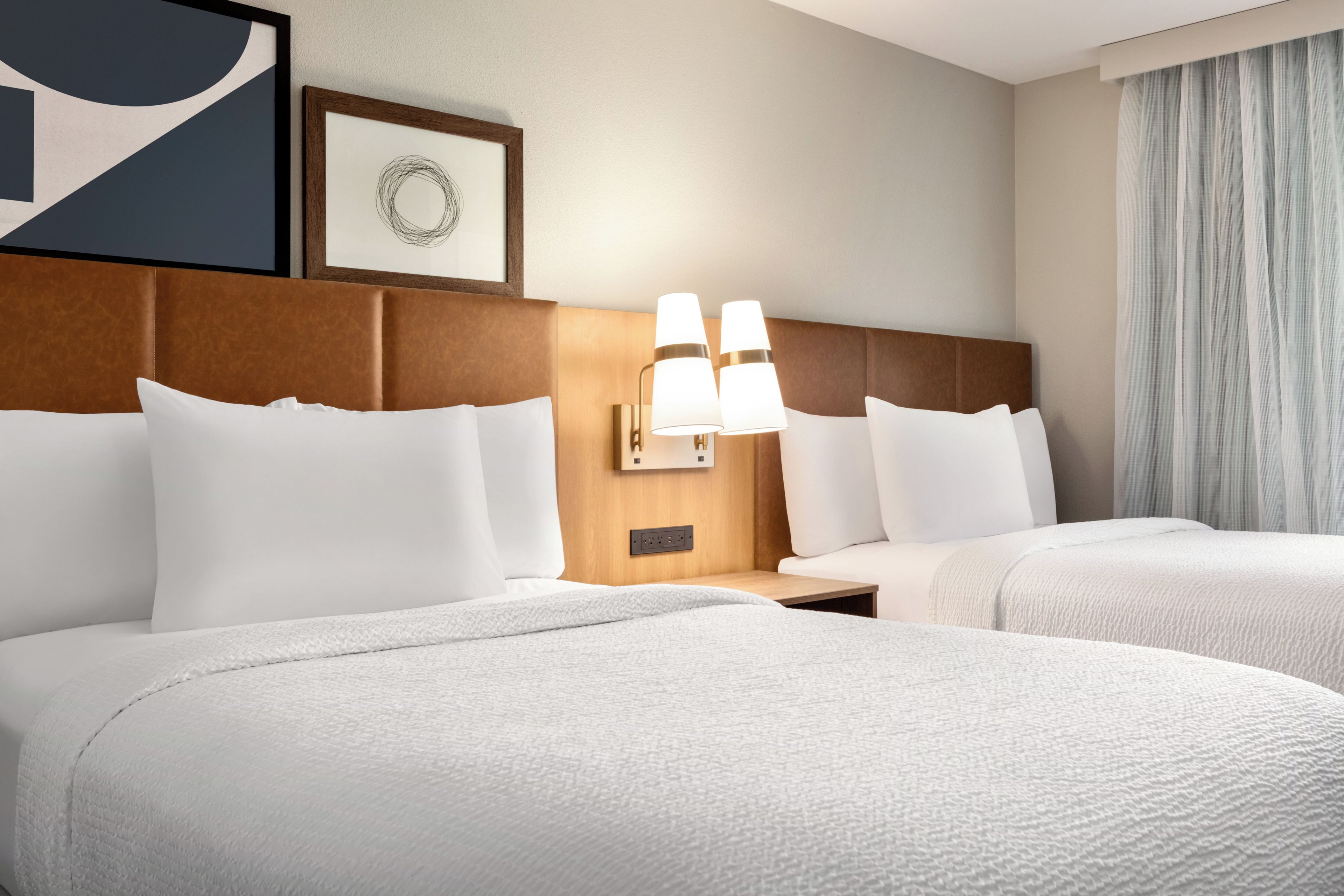 A hotel room with two beds, a headboard, bedside tables, lamps, and a framed picture on the wall.