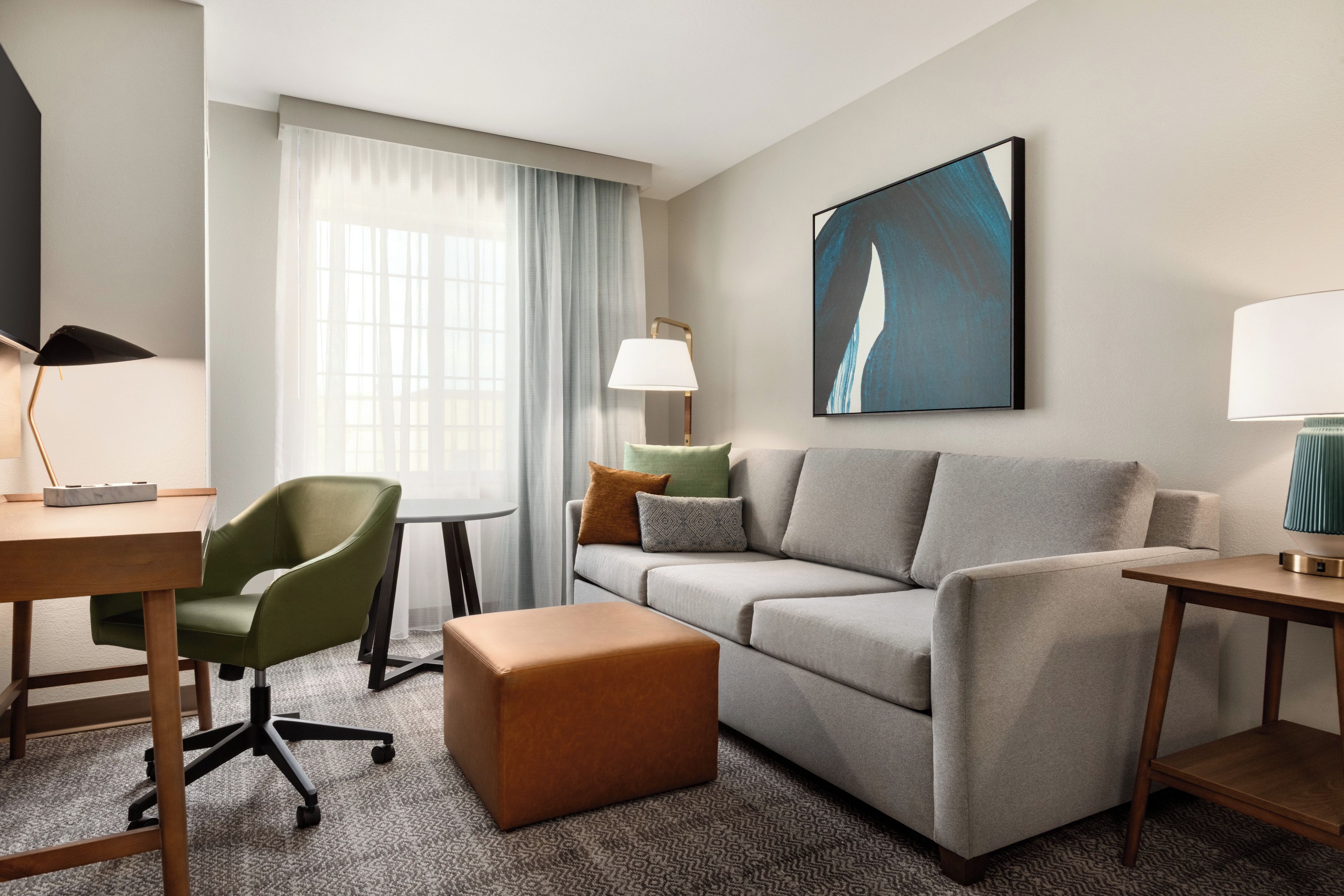 Una habitación de hotel moderna con un sofá gris, un sillón verde, una mesita de madera, una lámpara y un cuadro en la pared.