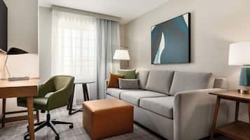 Una habitación de hotel moderna con un sofá gris, un sillón verde, una mesita de madera, una lámpara y un cuadro en la pared.