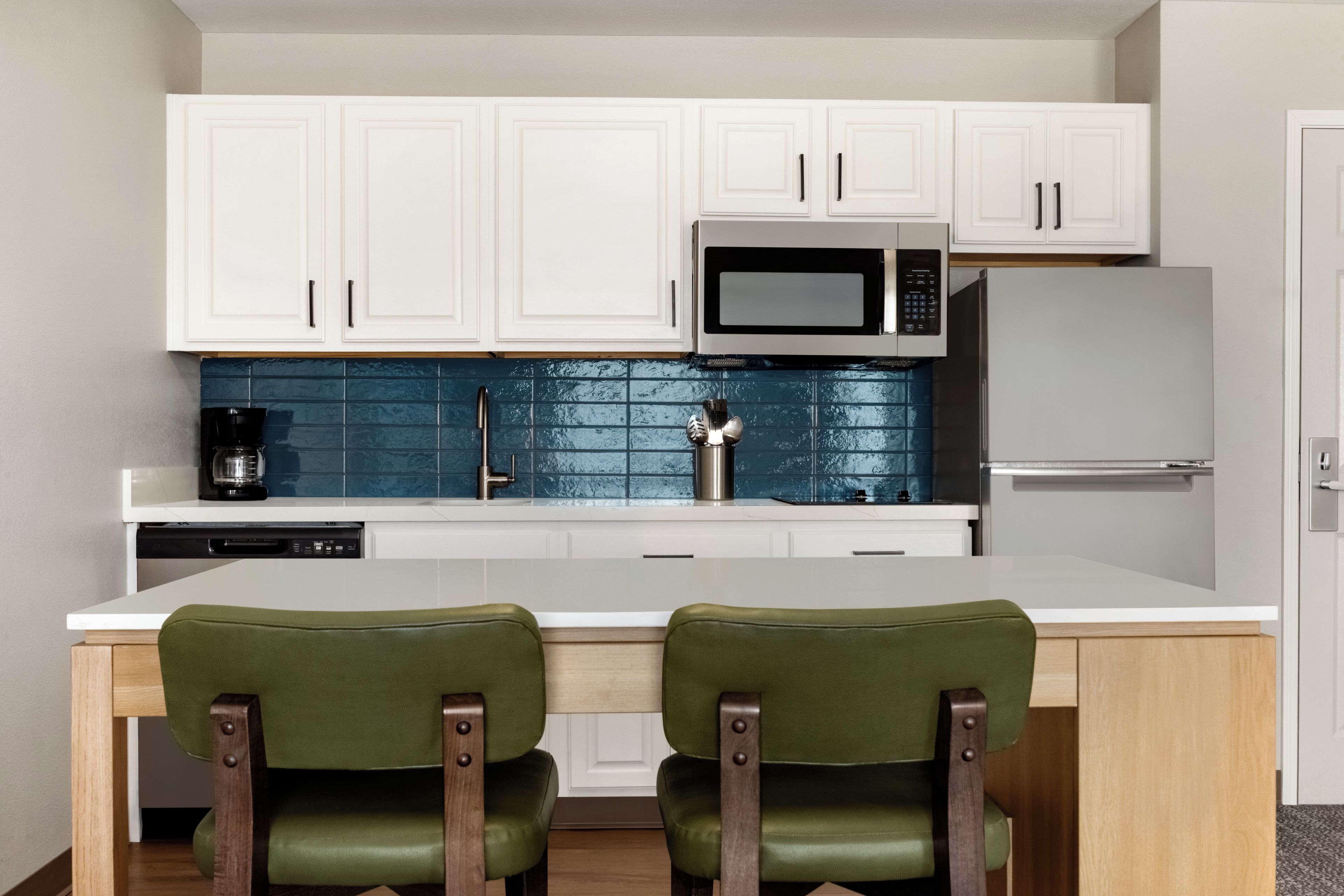 A modern kitchen with white cabinets, a microwave, a refrigerator, and a coffee maker. There are two green chairs at a white table.