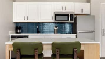 Una cocina moderna con armarios blancos, microondas, refrigerador y cafetera. Hay dos sillas verdes a una mesa blanca.