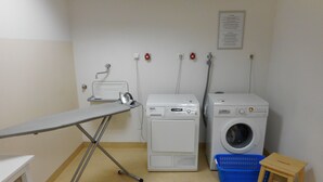 A laundry area with an ironing board, two washing machines, a blue laundry basket, and a small wooden stool. - Best Western Hotel Bamberg (Bamberg)