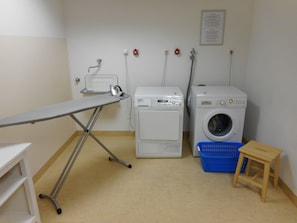 A laundry area with an ironing board, two washing machines, a blue laundry basket, and a small wooden stool. - Best Western Hotel Bamberg (Bamberg)
