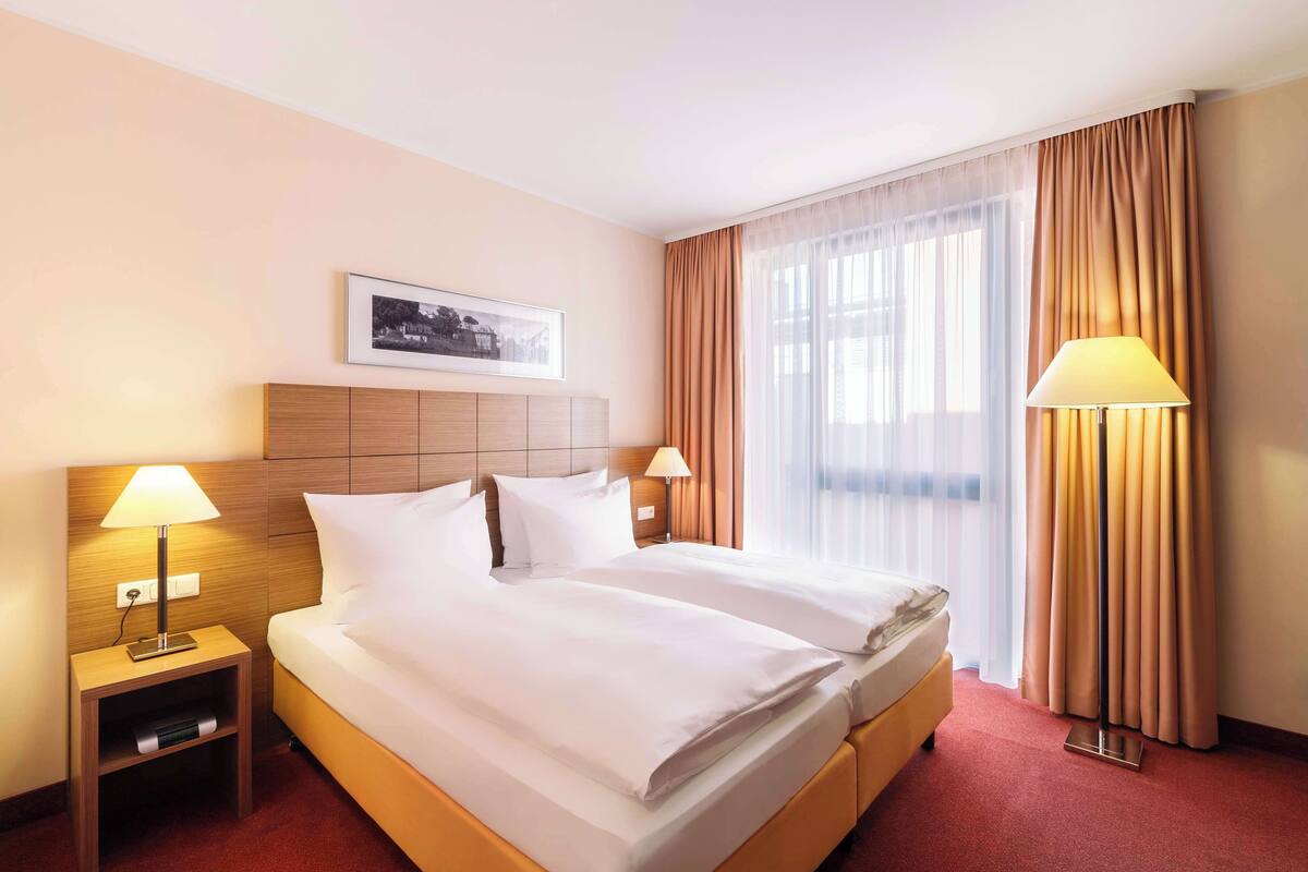 A hotel room with a double bed, two bedside lamps, a nightstand, a framed picture on the wall, and a window with curtains.