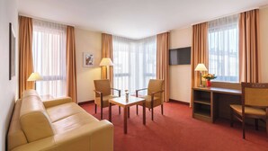 A hotel room with a sofa, two chairs, a small table, a TV, and a desk with a lamp and flower vase. - Best Western Hotel Bamberg (Bamberg)