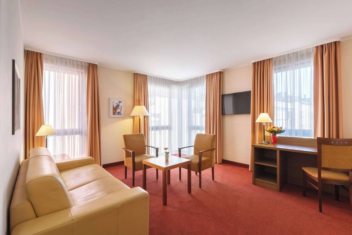A hotel room with a sofa, two chairs, a small table, a TV, and a desk with a lamp and flower vase.