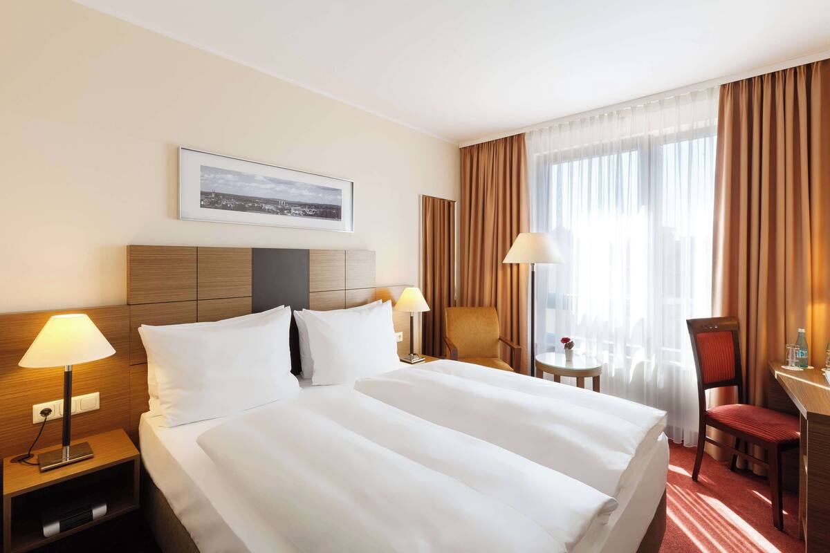 A hotel room with a large bed, two bedside tables, a desk, a chair, and a window with curtains.