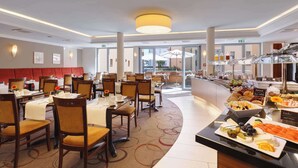 Un comedor de hotel con varias mesas y sillas, un bufé con diversas opciones de comida y vistas al exterior a través de amplios ventanales.