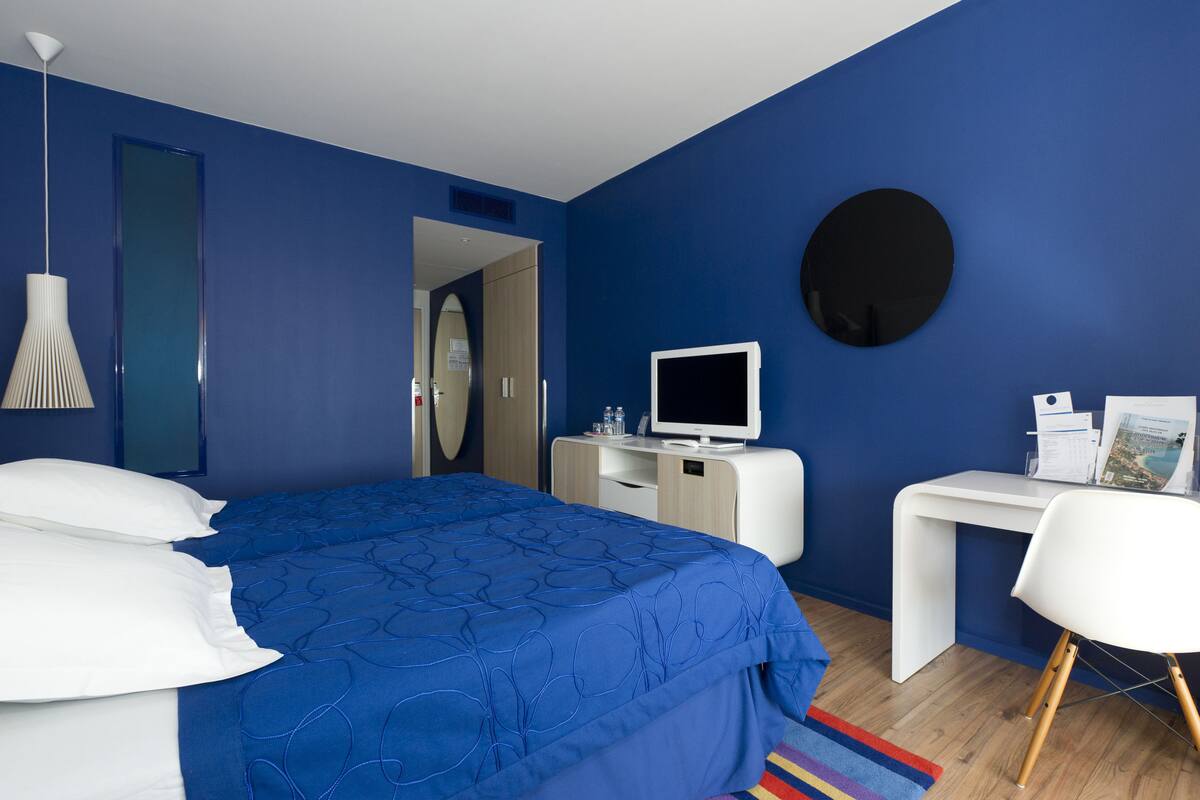 Une chambre d’hôtel moderne, dotée d’un grand lit, d’un bureau, d’une télévision et d’un grand miroir rond accroché au mur.